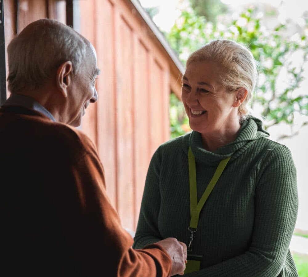 A younger female carer entering the door smiling while shaking hand with an older male adult
