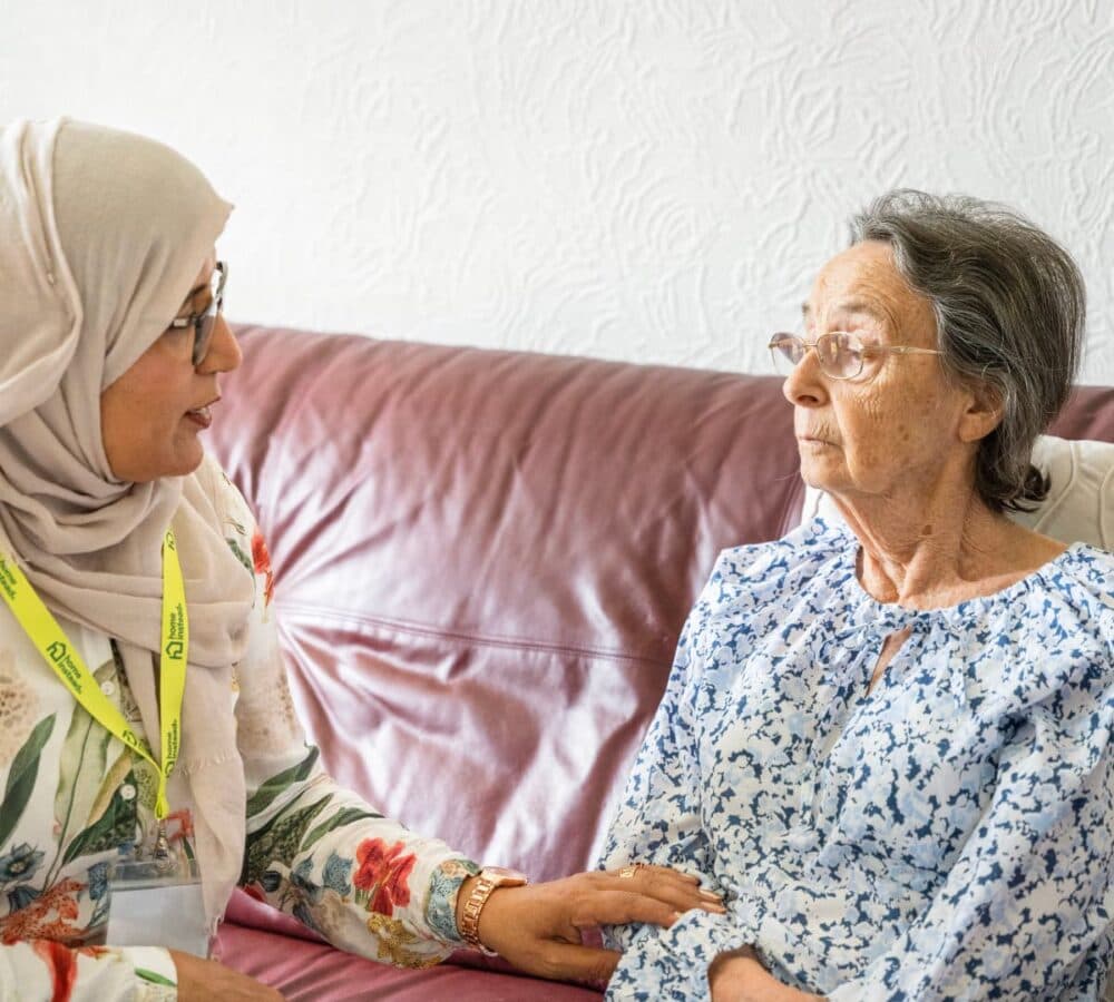 A female carer wearing eyeglasses while sitting on a couch and talking to an older female adult with grey hair and sitting on a couch