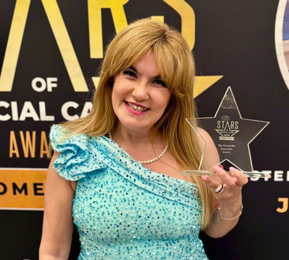 Smiling woman in a blue dress holds a star-shaped award in front of a "Stars of Social Care" backdrop. - Home Instead