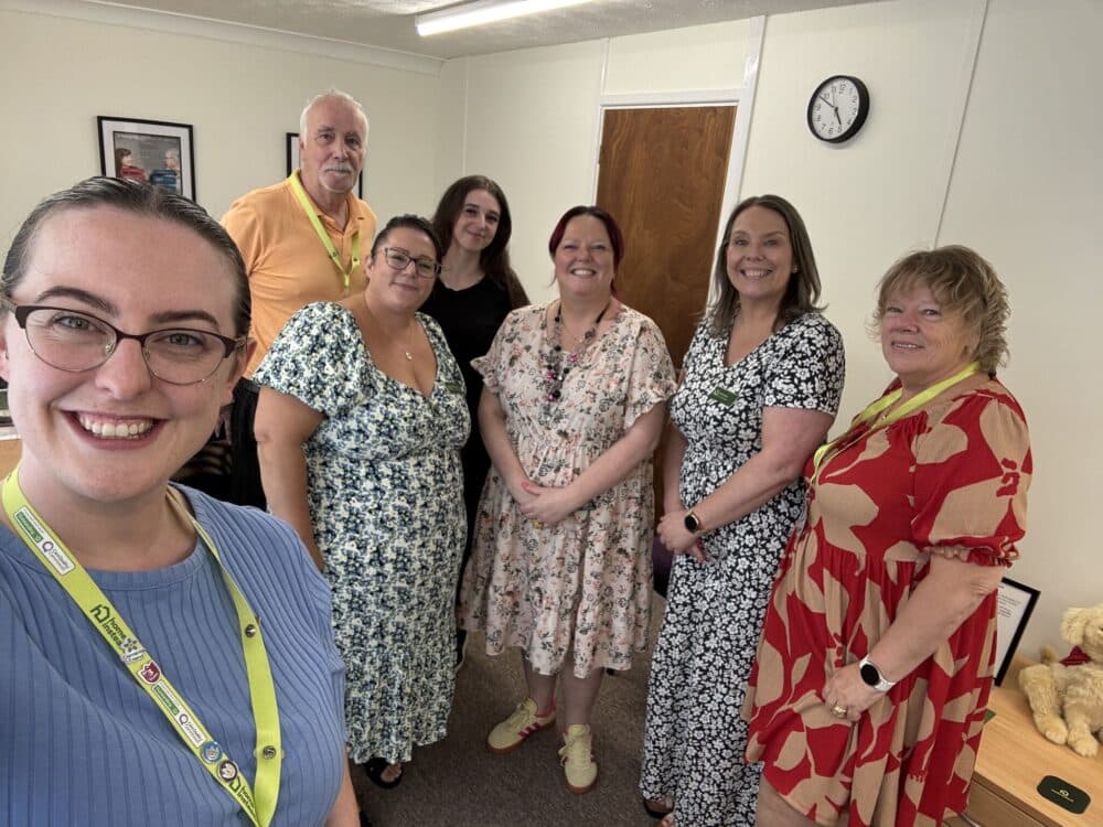 Seven adults smiling and posing together in an office with a clock and door in the background. - Home Instead