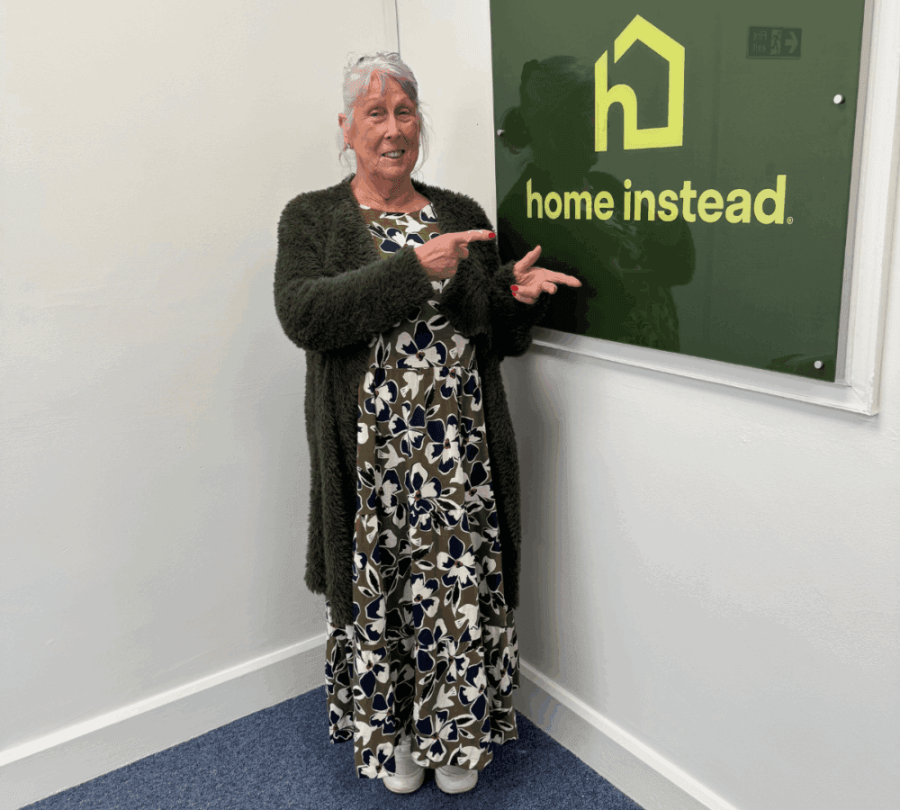 Smiling woman with purple hair points at a "home instead" sign on a wall in an office setting. - Home Instead