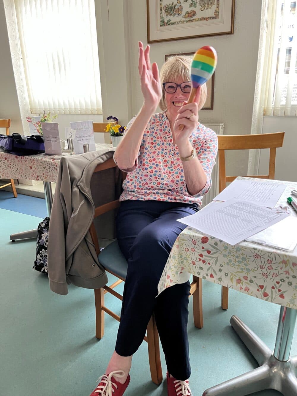 Smiling woman sitting, holding a rainbow maraca, clapping in a bright room with tables and papers. - Home Instead