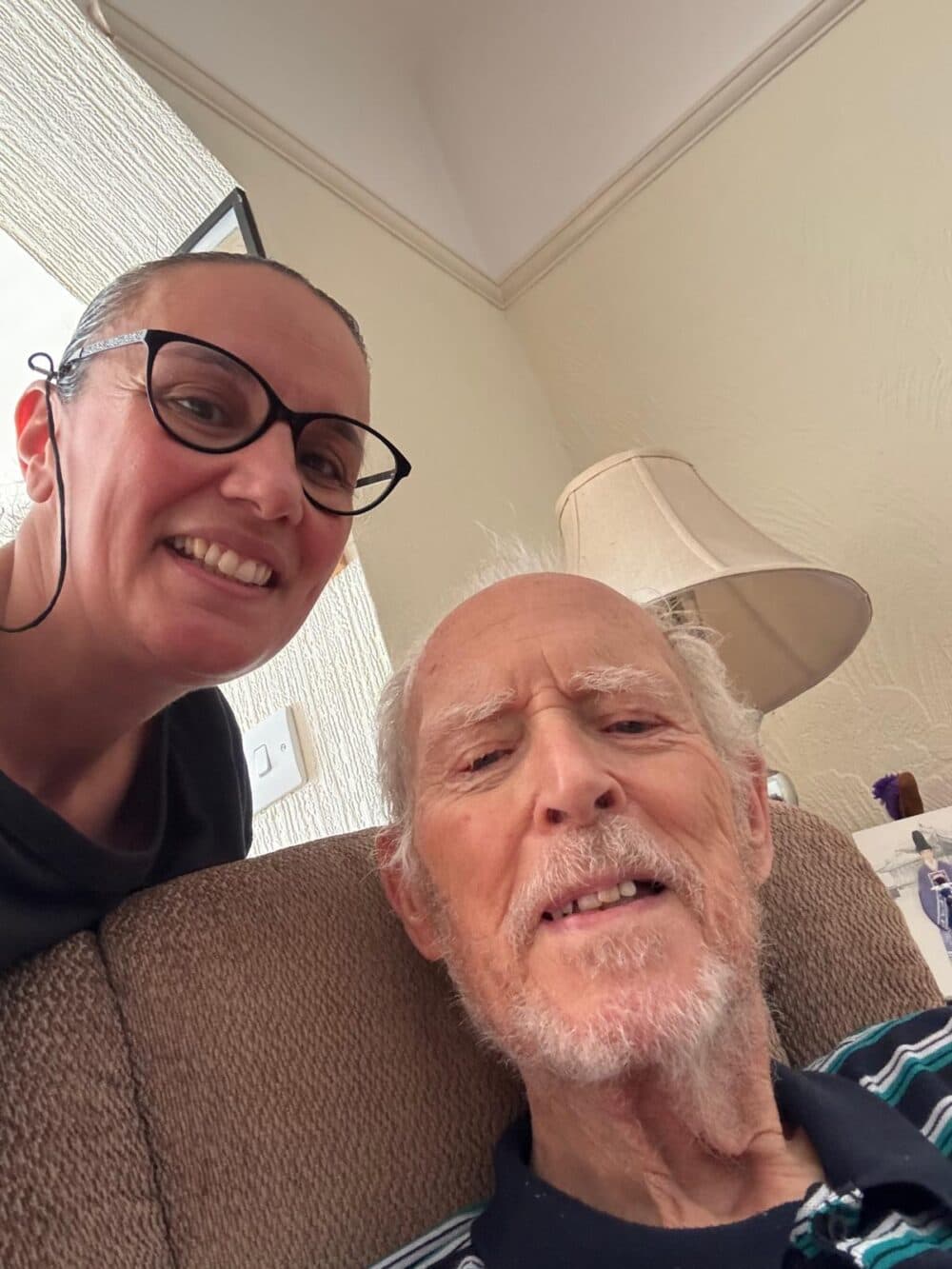 Smiling woman in glasses takes a selfie with an elderly man sitting on a chair in a cozy room. - Home Instead