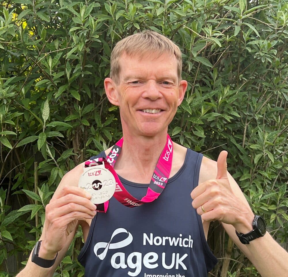 Smiling man in a "Norwich Age UK" vest holds a race medal and gives a thumbs up in front of green foliage. - Home Instead