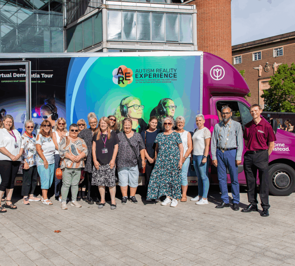 A group of people stand smiling in front of an Autism Reality Experience van on a sunny day. - Home Instead