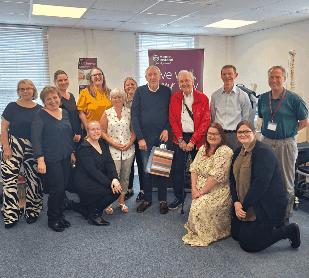 A group of people smiling, standing and kneeling together in an office with a Home Instead banner behind them. - Home Instead
