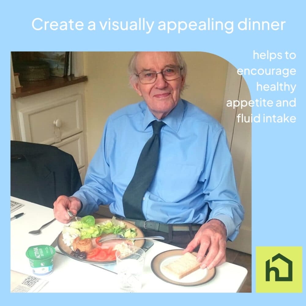 An elderly man in a blue shirt smiles at dinner, with salad, bread, and a drink on the table in front of him. - Home Instead