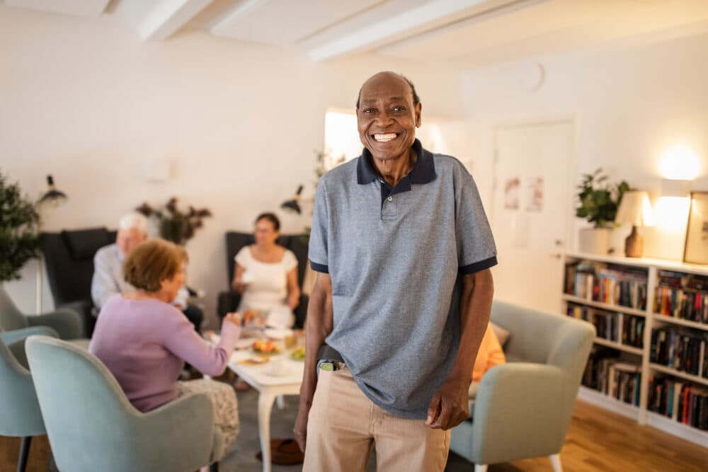 Smiling man stands in a cozy living room while three people sit and talk at a table in the background. - Home Instead
