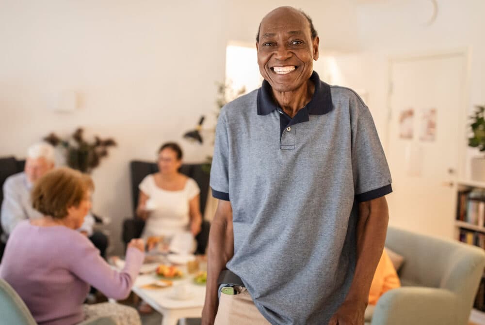 Smiling man stands in a living room while others chat and eat at a table in the background. - Home Instead