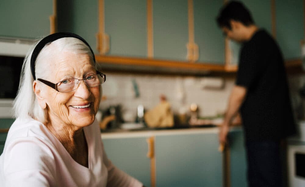 Smiling elderly woman with glasses sits in kitchen; a man stands in the background near the counter. - Home Instead