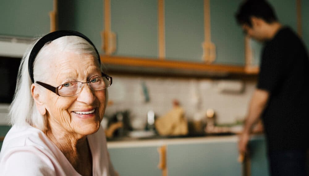 Smiling elderly woman with glasses in foreground; person standing in kitchen background. - Home Instead