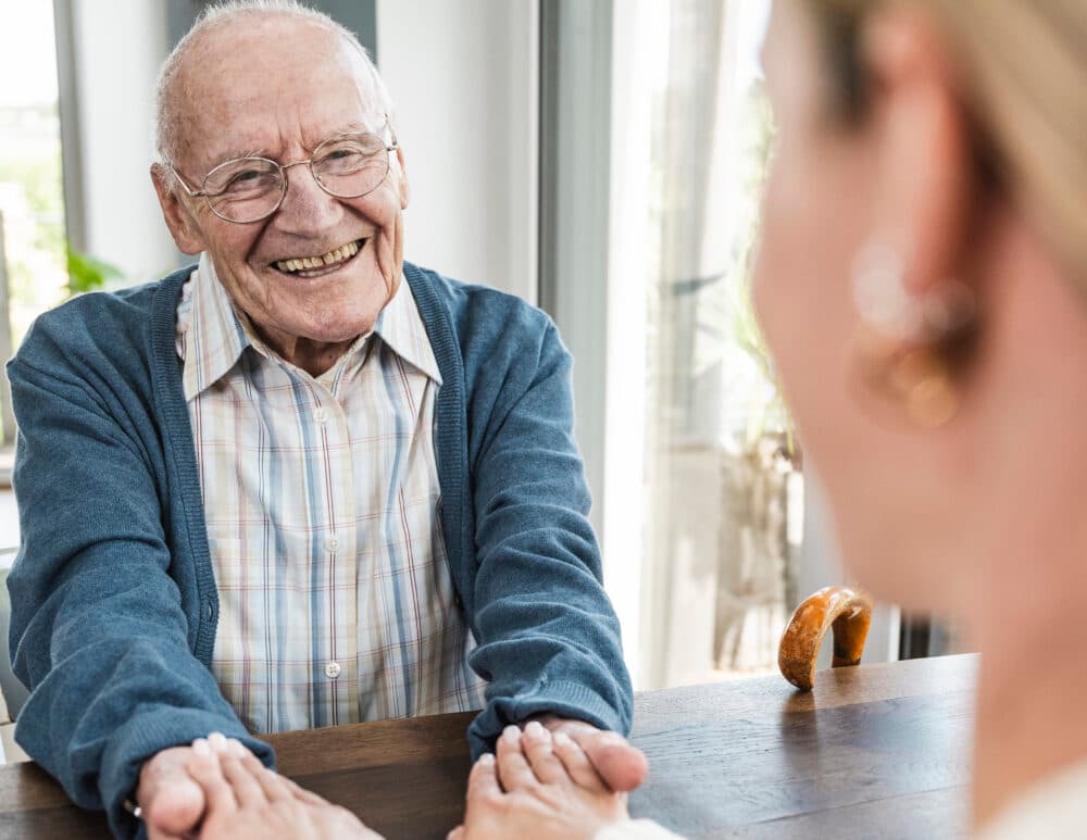Smiling elderly man holding hands with a woman across a table in a bright room. - Home Instead
