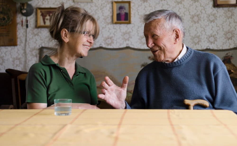 Smiling woman and elderly man having a friendly conversation at a table in a cozy room. - Home Instead