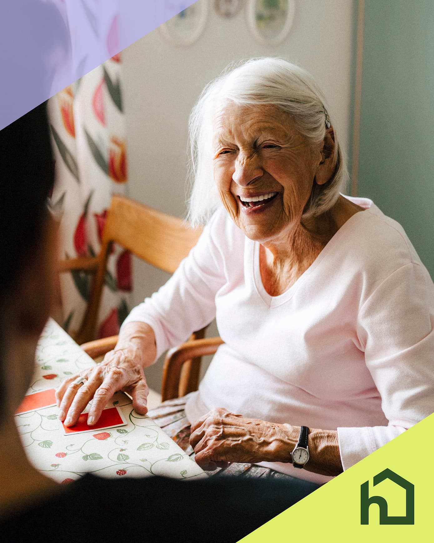 Smiling elderly woman sitting at a table, chatting with someone, with a green home icon in the corner. - Home Instead
