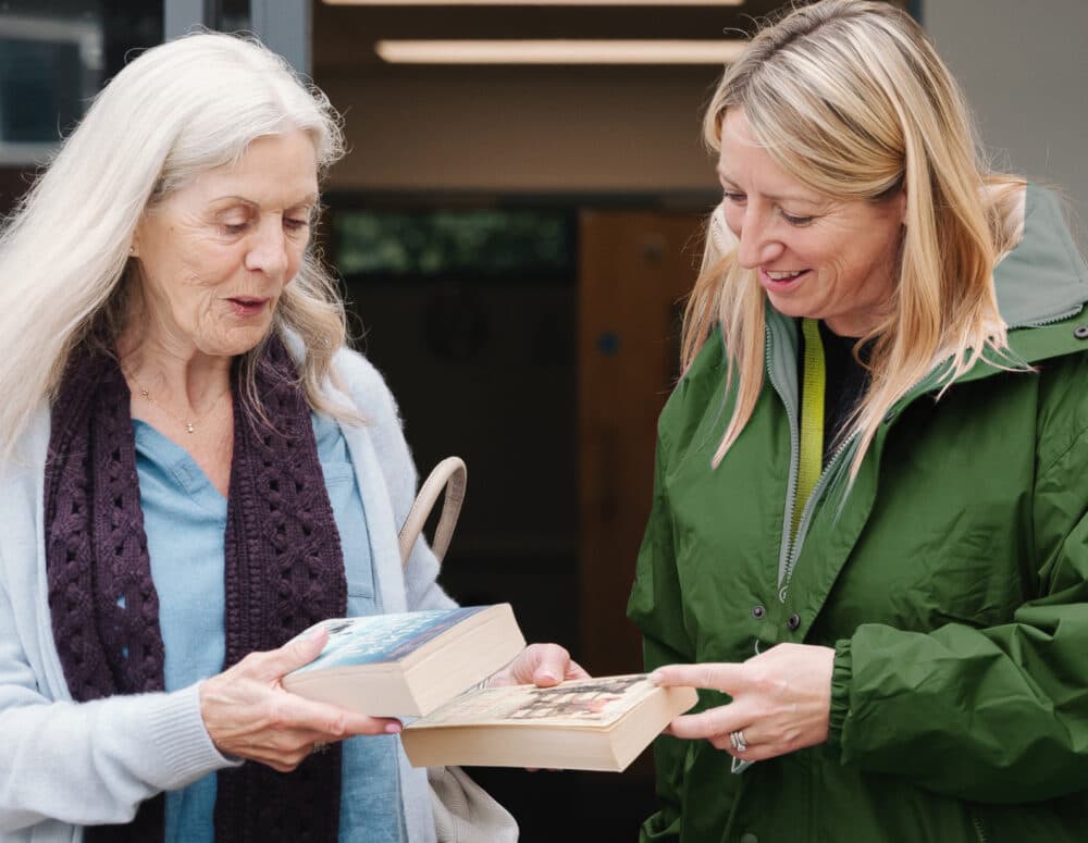 Two women standing together, smiling and exchanging books outside a building. - Home Instead