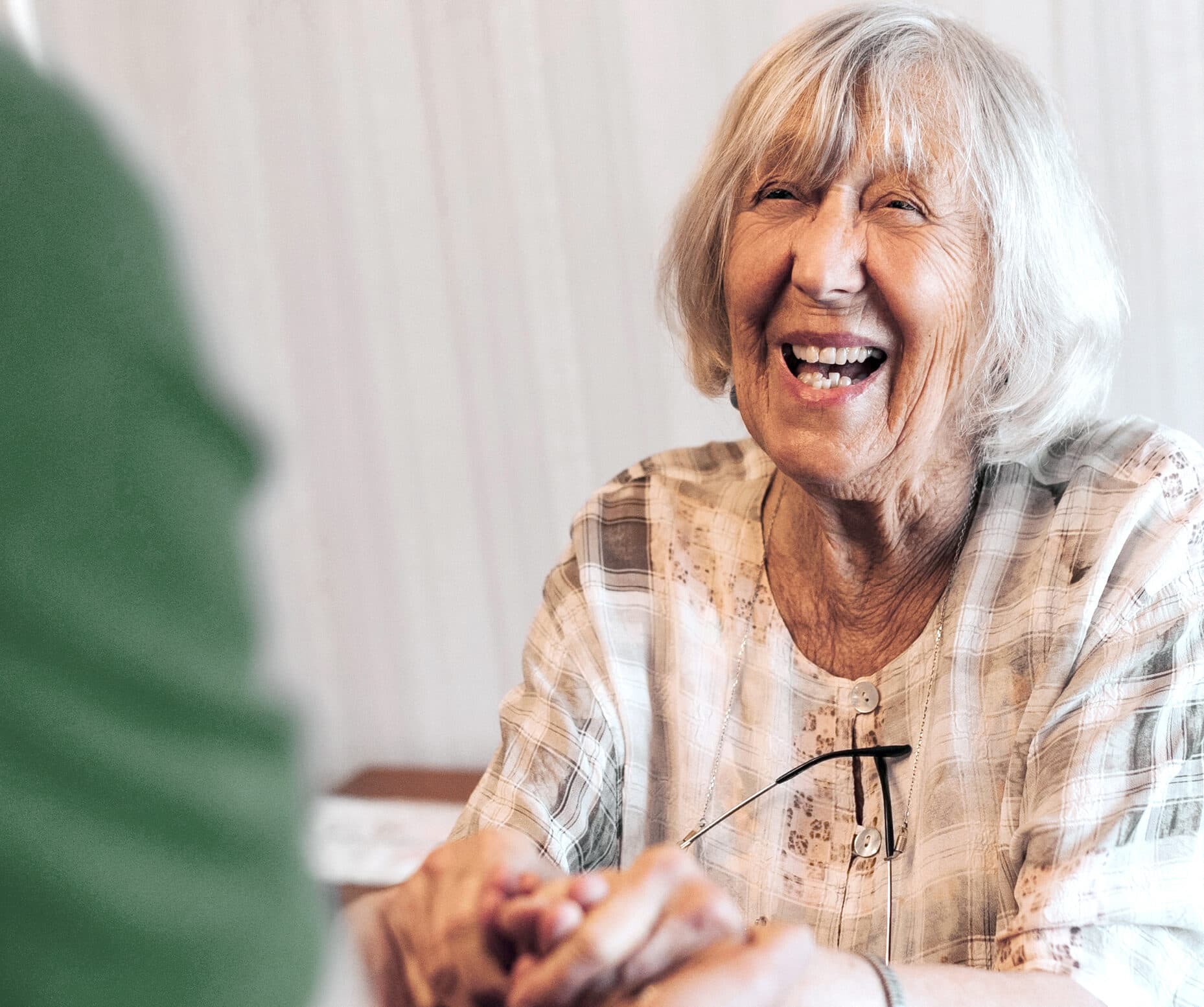 Smiling elderly woman holds hands with someone, wearing a light plaid blouse and glasses around her neck. - Home Instead
