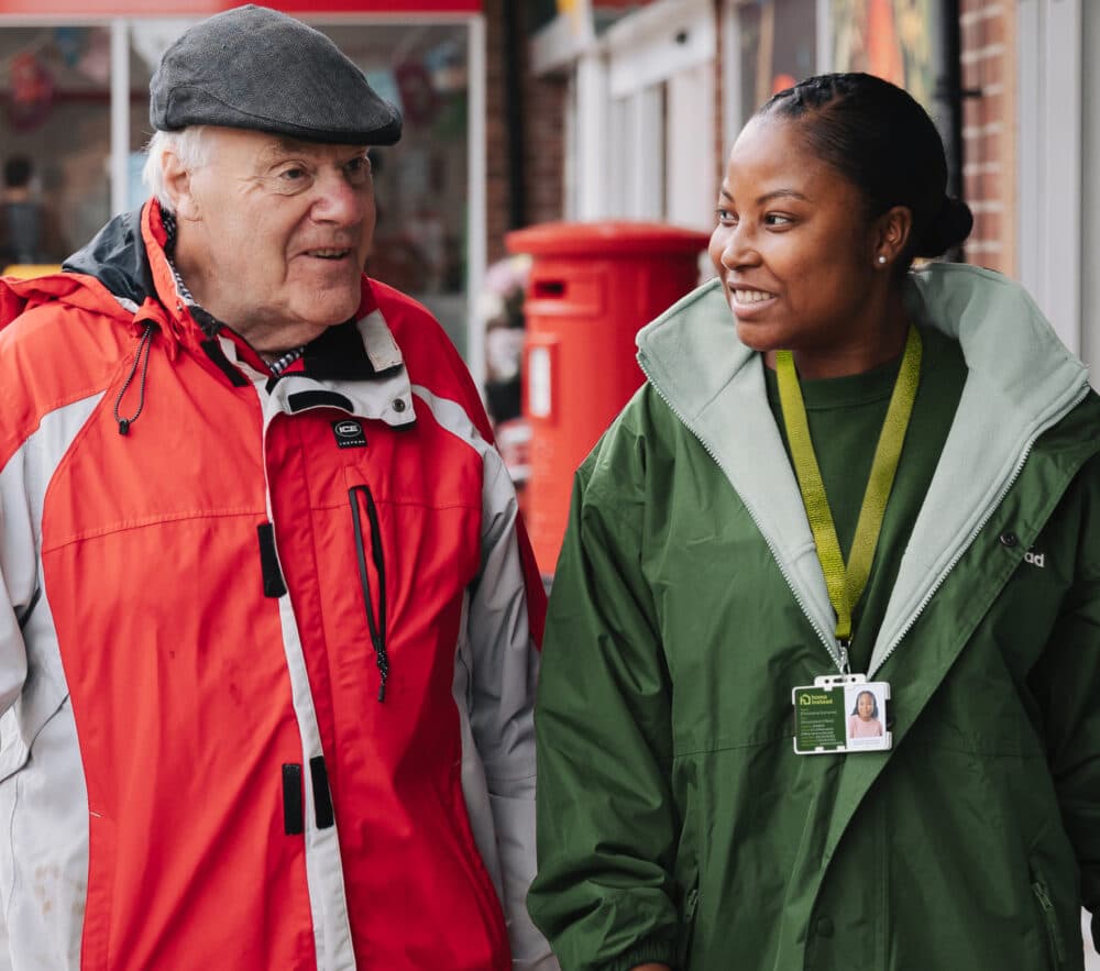 An elderly man and a woman in a green jacket smiling and walking together outside. - Home Instead