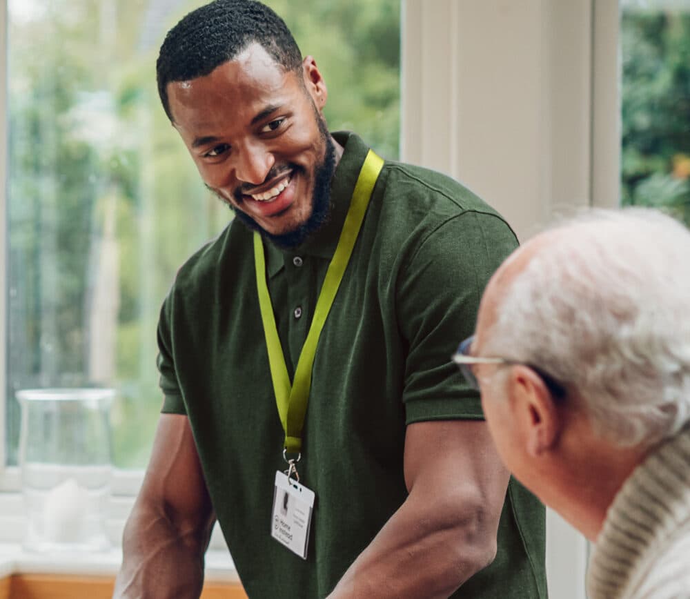 A smiling caregiver with a name badge talks to an elderly man in a bright room. - Home Instead
