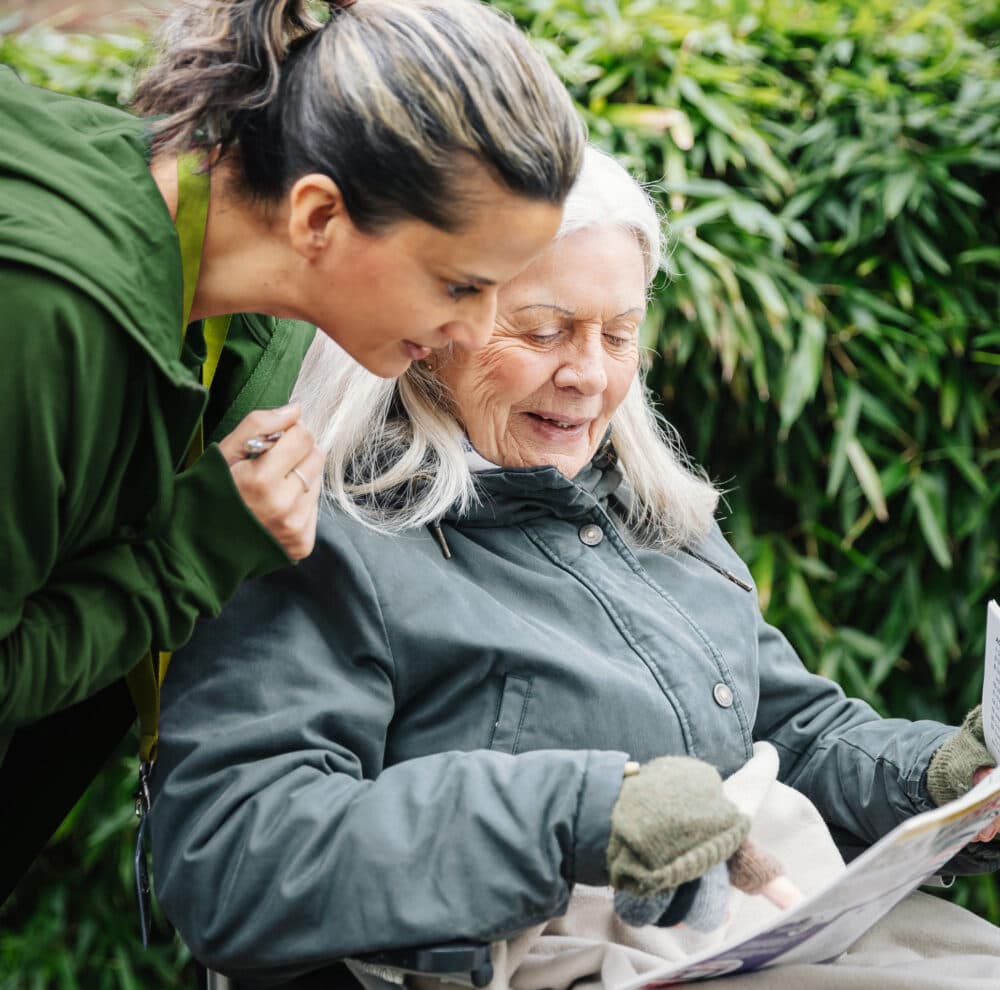 A younger woman and an older woman smile while reading a paper together outdoors. - Home Instead