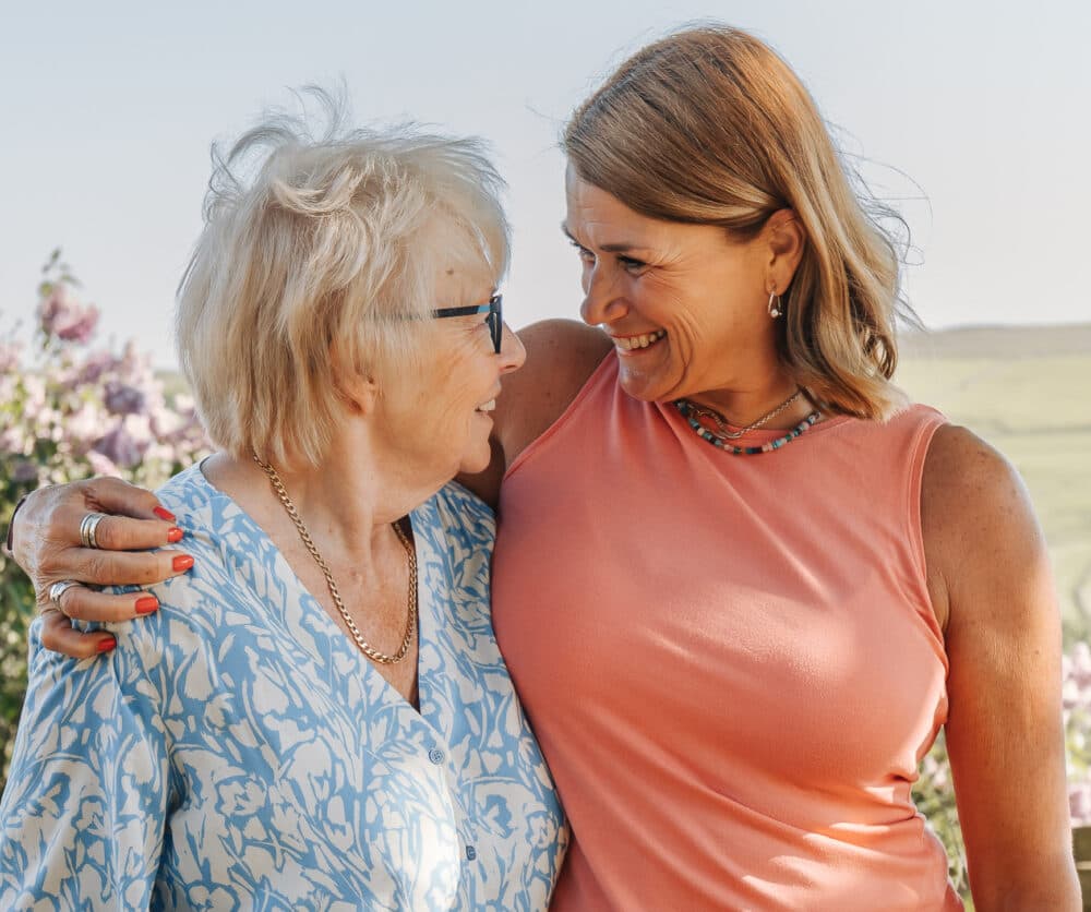 Two women smiling at each other outdoors, one older and one younger, with arms around each other. - Home Instead