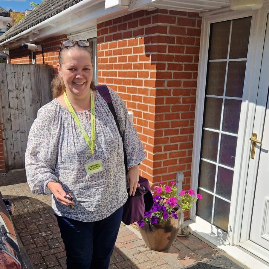 Smiling woman with a lanyard stands outside a brick house next to a white door and a pot of flowers. - Home Instead