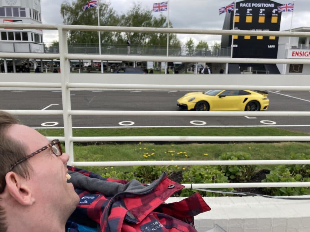 A person smiling in the foreground with a yellow sports car driving on a racetrack in the background. - Home Instead