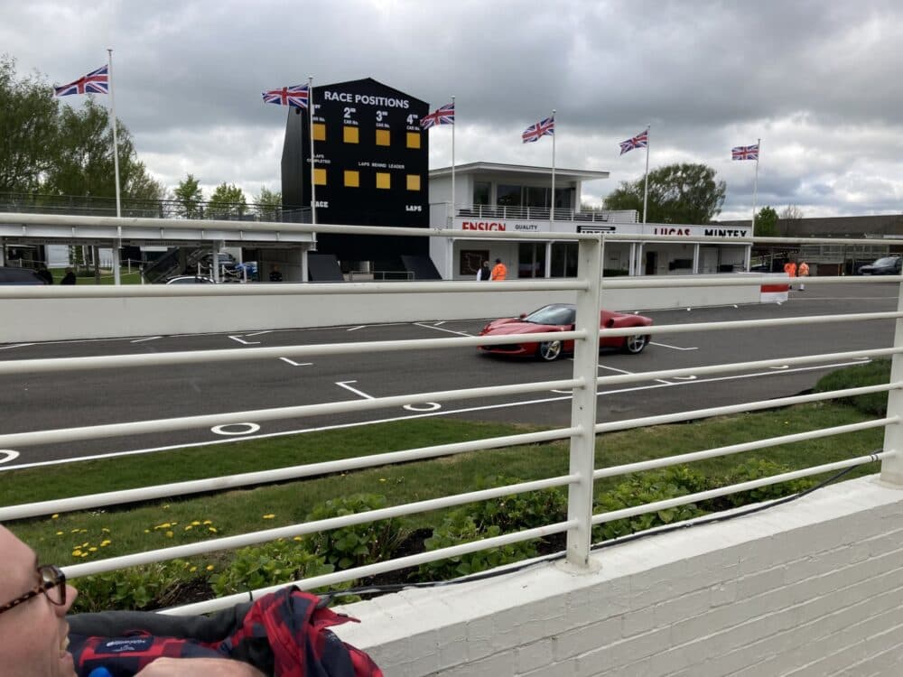 A red sports car drives past a race scoreboard on a cloudy day at a racetrack, seen through a white fence. - Home Instead