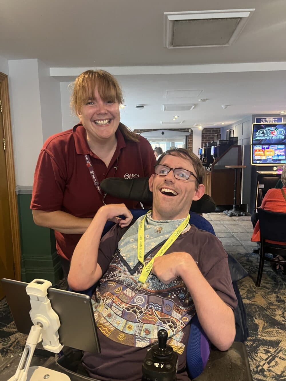 A smiling woman stands behind a man in a wheelchair, both looking happy in a warmly lit room. - Home Instead