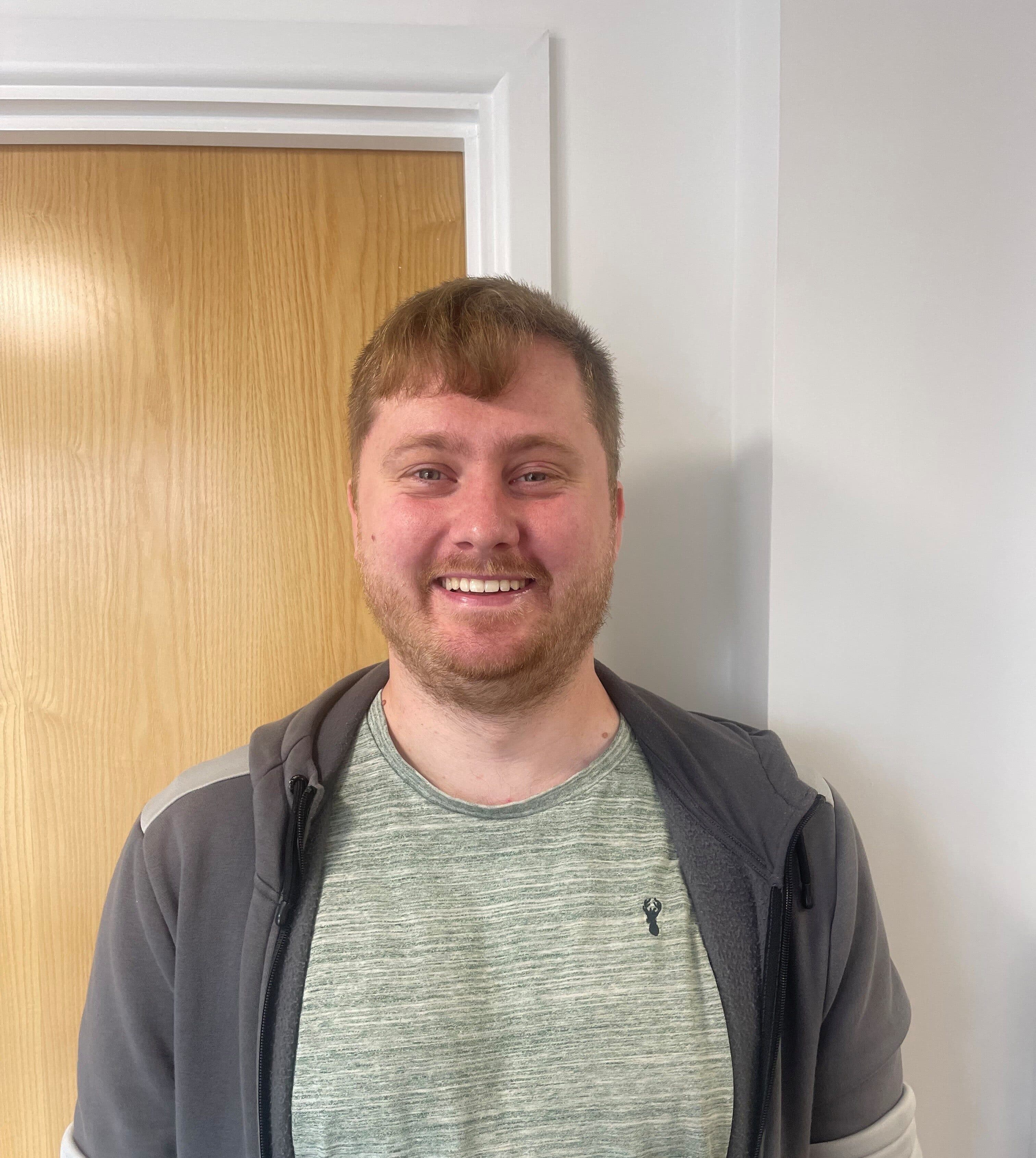A man smiling while standing indoors in front of a wooden door. He is wearing a light green T-shirt and a gray zip-up hoodie with the sleeves rolled up.
