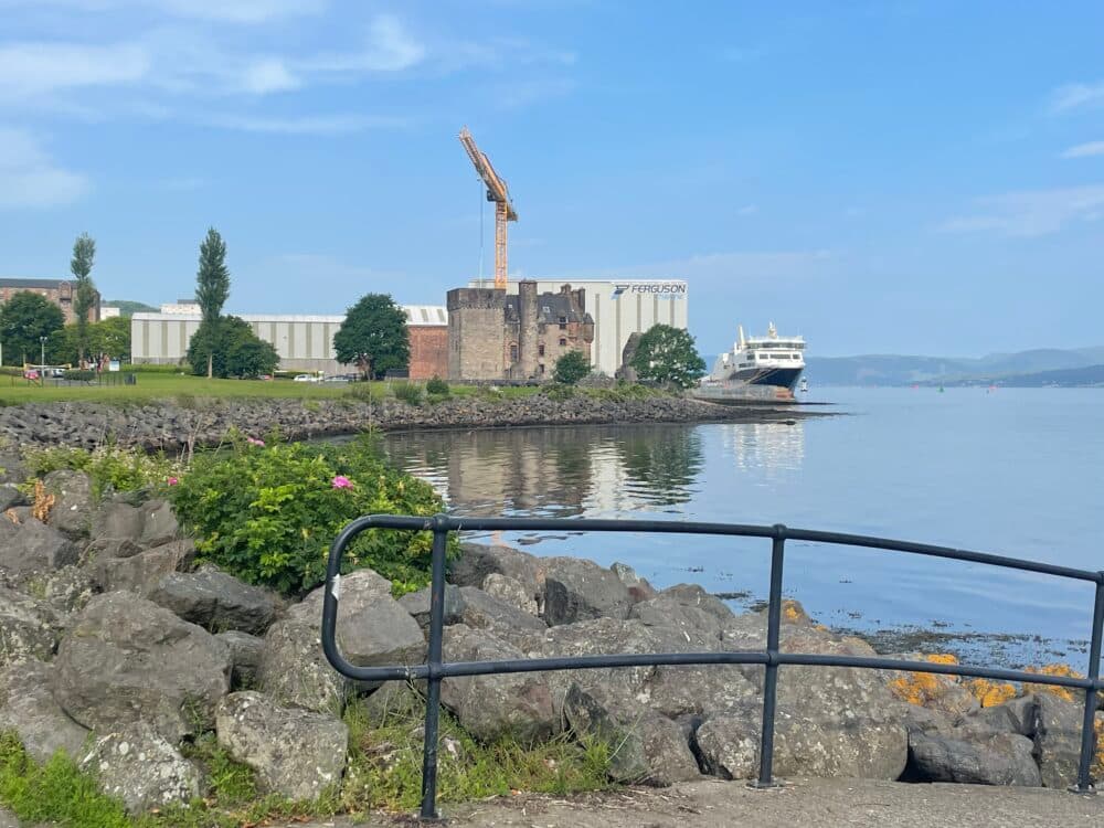 Rocky shoreline with a black rail, ship docked near a castle, and a crane under a blue sky. - Home Instead