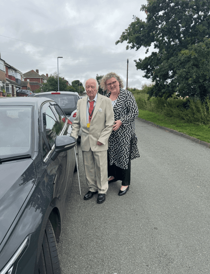 An elderly man in a suit and a woman in a patterned dress stand by a parked car on a suburban street. - Home Instead