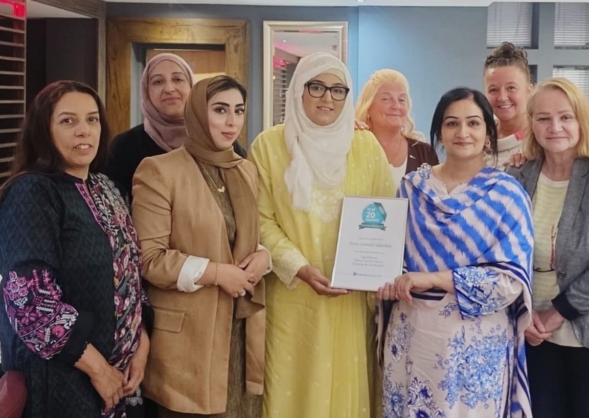A diverse group of women stand together, one holding a framed certificate, all smiling at the camera. - Home Instead