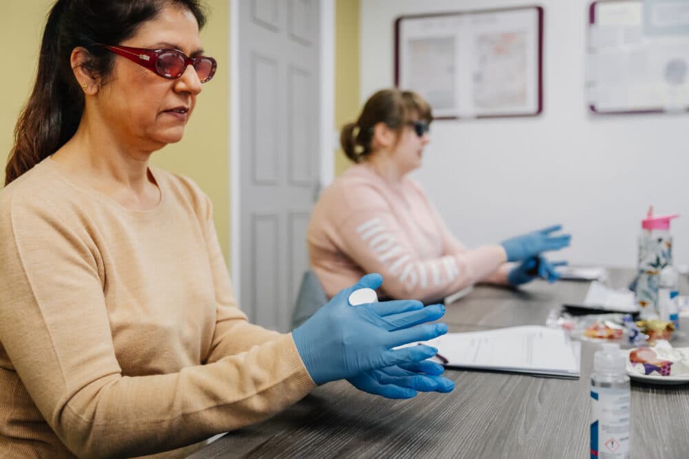 Two women wearing blue gloves and safety glasses sit at a table with papers and lab materials. - Home Instead