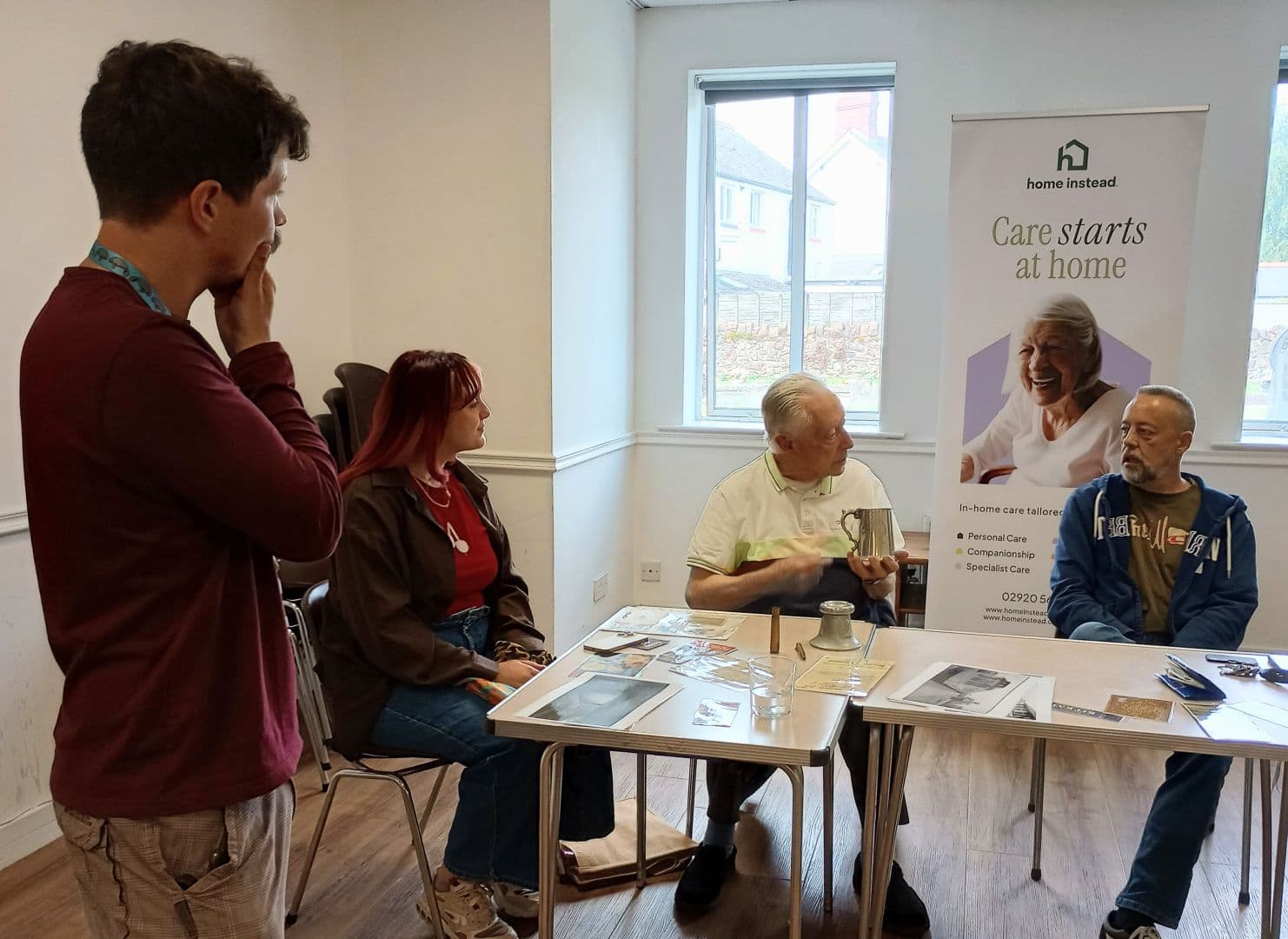Four people sit and talk around a table in a bright room with a "Care starts at home" banner in the background. - Home Instead