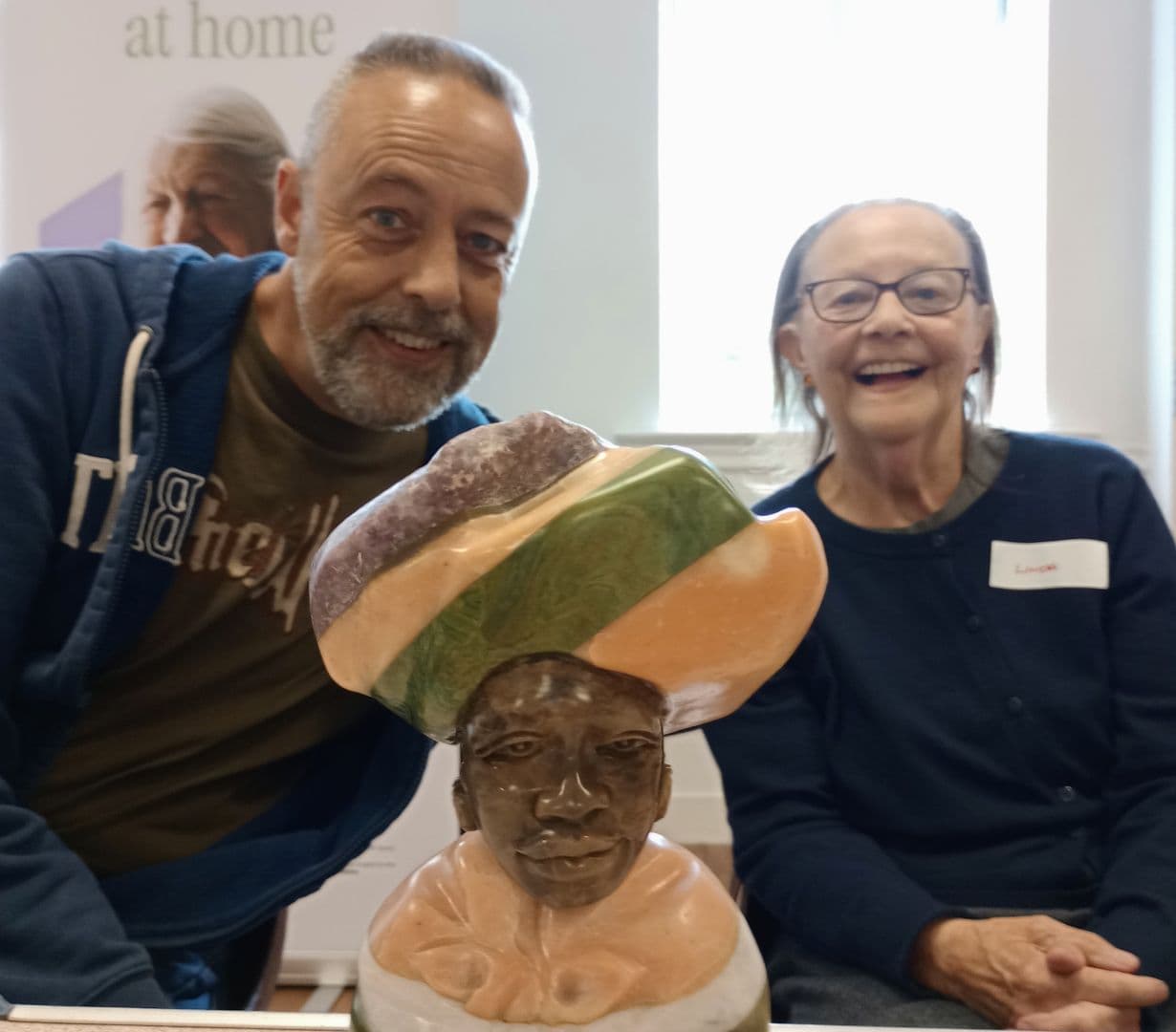 Two smiling people sitting behind a colorful bust sculpture, indoors with natural light from a window. - Home Instead