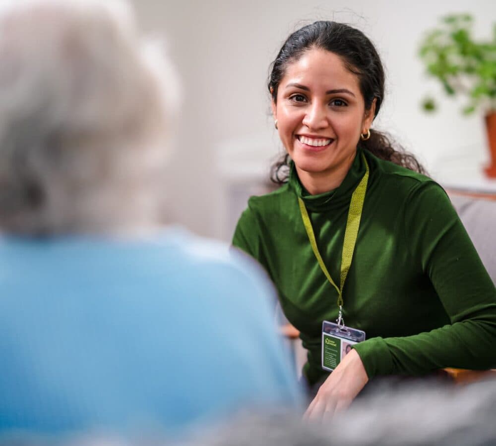 Smiling caregiver in a green shirt talks with an older person in a bright, comfortable room. - Home Instead