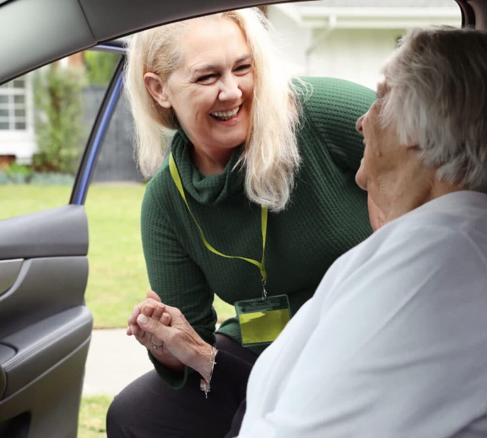 Smiling woman helps an older woman out of a car, holding her hand and making eye contact. - Home Instead