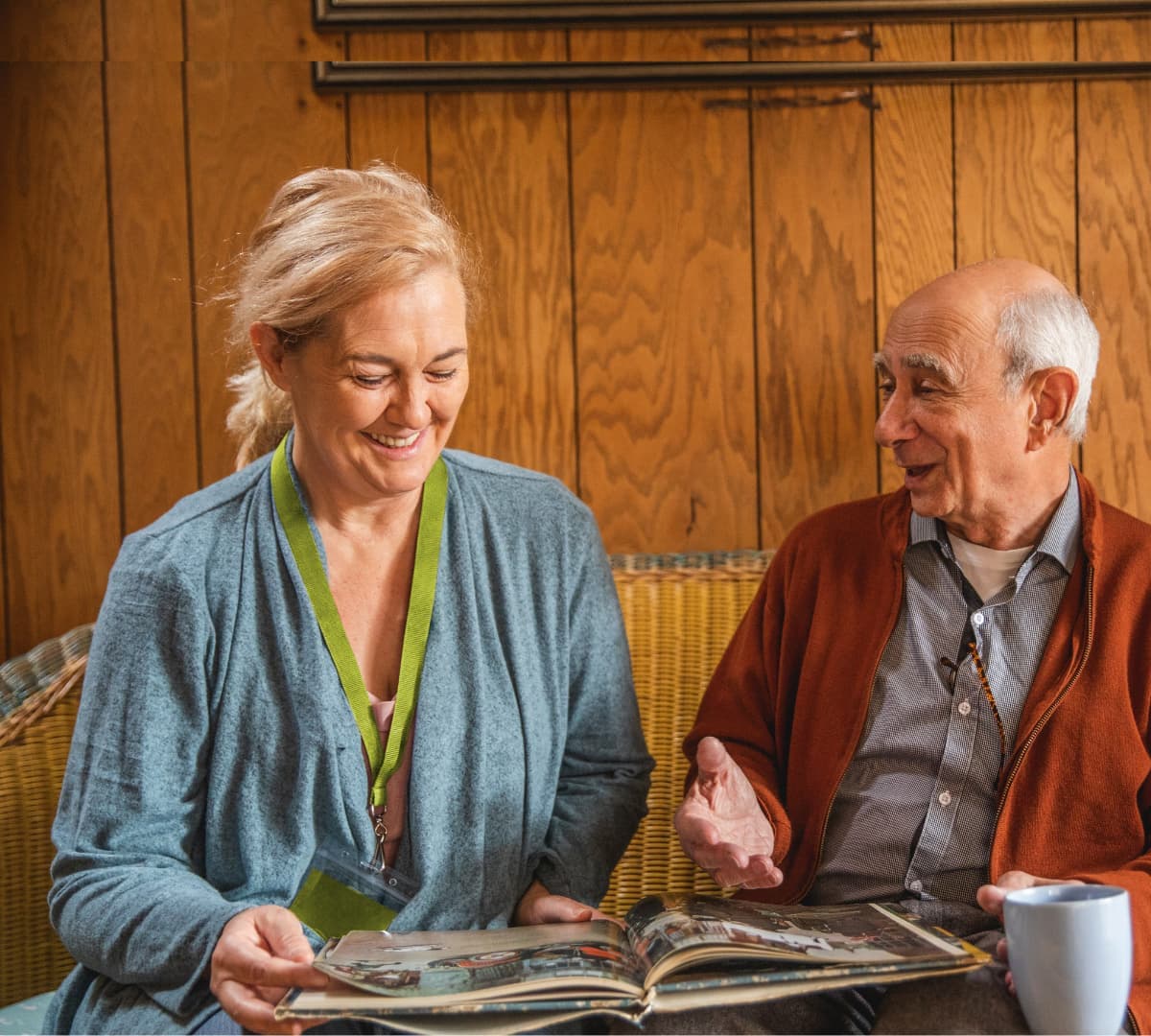 An older male adult with grey hair looking at a photo album while sitting on a couch with his younger female carer with blonde hair and both are happy and smiling