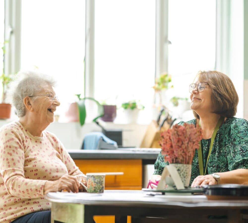 Two women chatting inside the kitchen while drinking coffee both happy and smiling