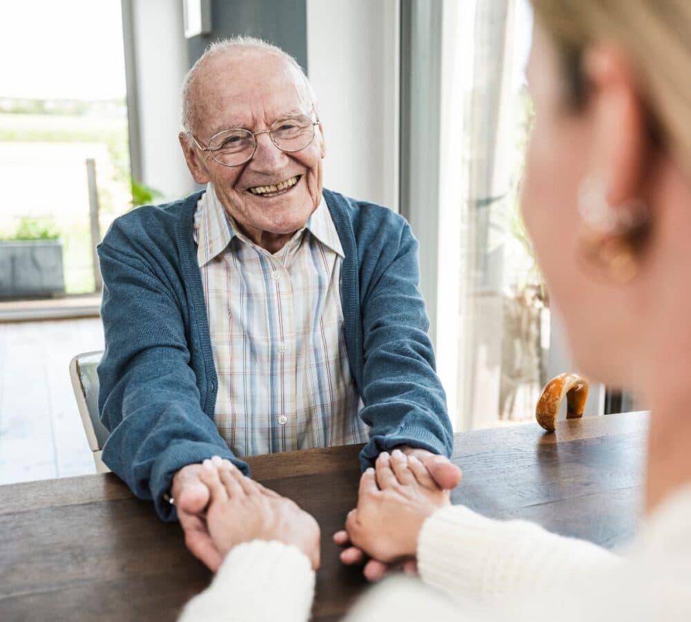 An older male adult wearing blue sweater happy and smiling while sitting on a chair and holding the hand of his younger female carer