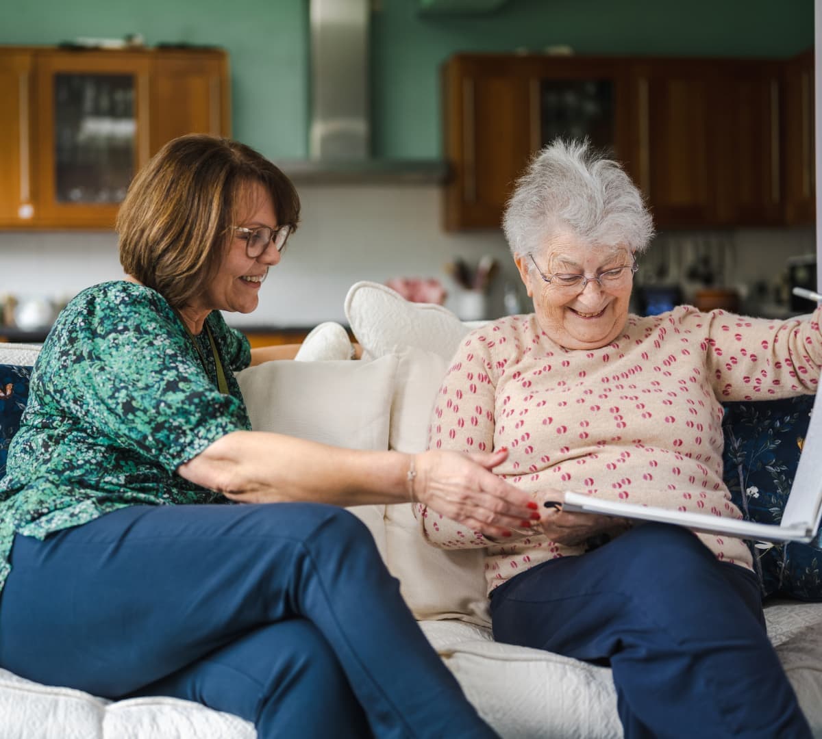 Two older women sit on a couch, smiling and looking at a photo album together in a cozy kitchen. - Home Instead