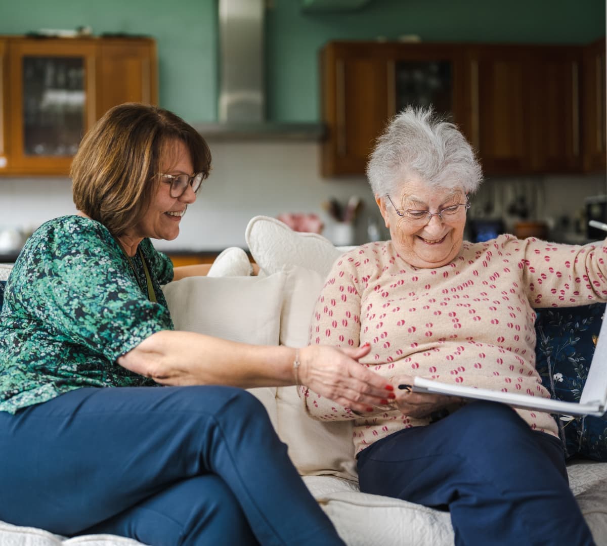 Two women looking at an album while sitting on a couch inside the house both happy and smiling