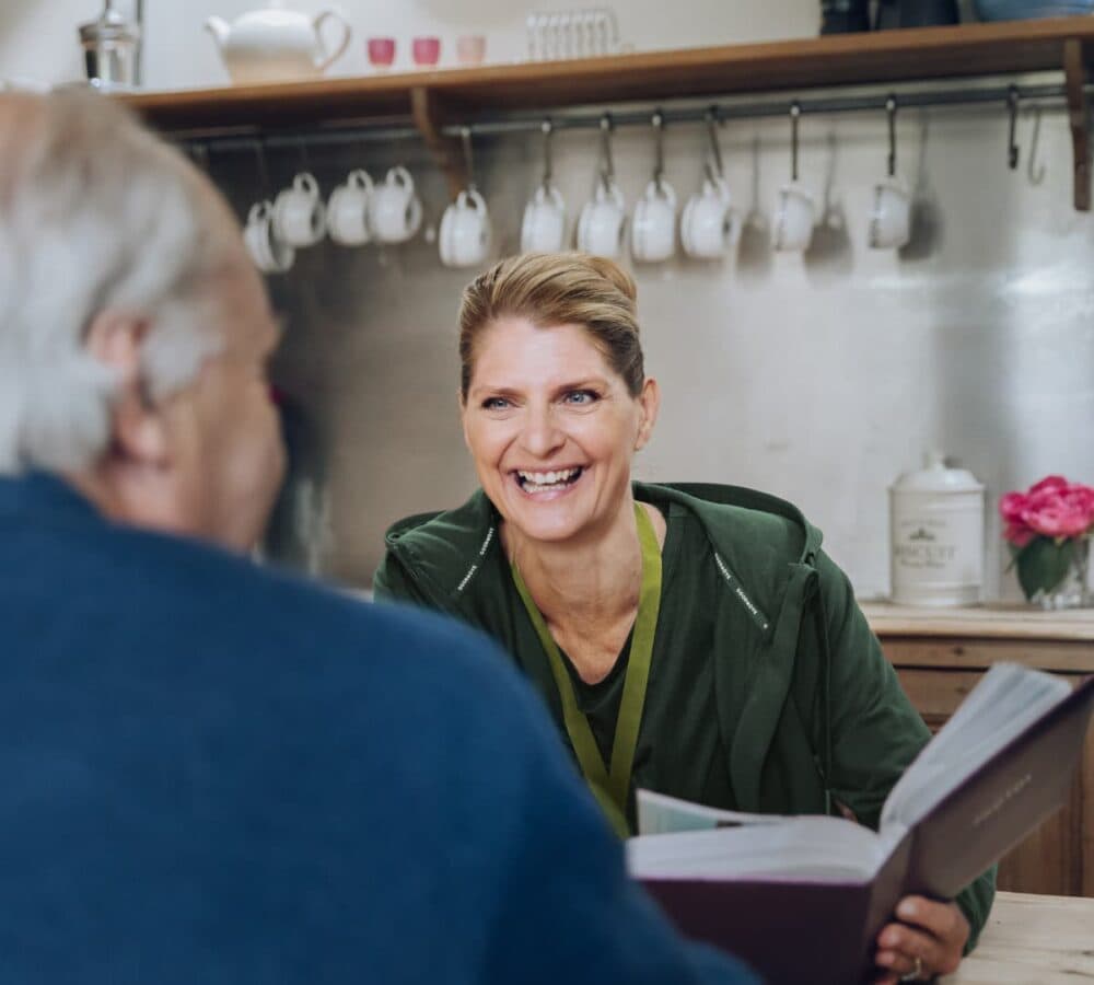 A young female adult with blonde hair and wearing green sweatshit chatting with an olde male adult while holding a photo album insode the kitchen