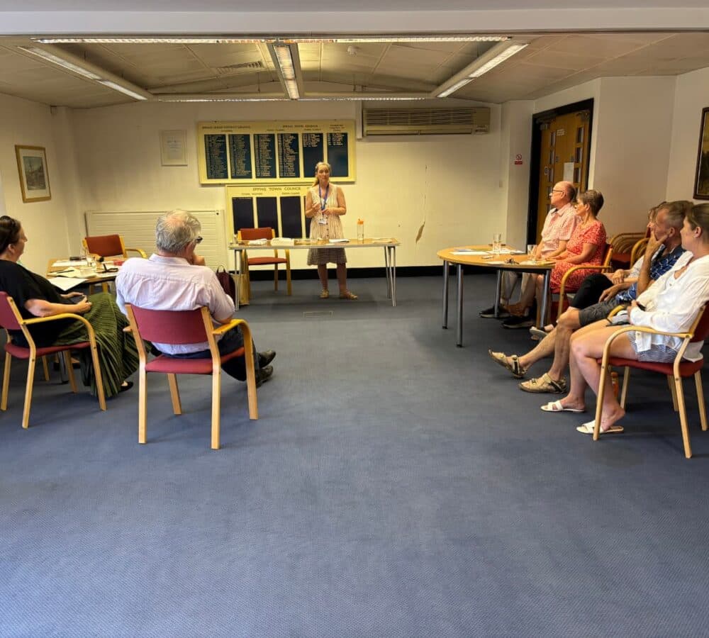 A woman stands and speaks to a group of seated people in a meeting room with blue carpet. - Home Instead