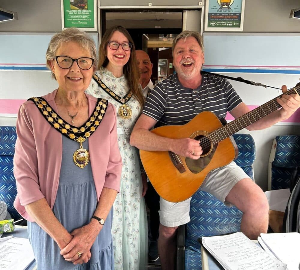 Four smiling adults pose on a train; one man plays guitar, two women wear ceremonial chains and dresses. - Home Instead