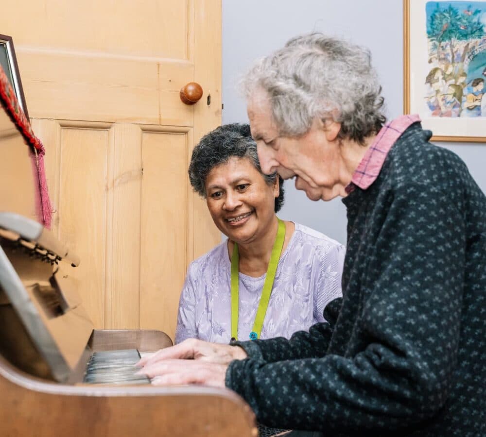 An older male adult with grey hair playing the piano while his younger female carer is sitting beside her and listening