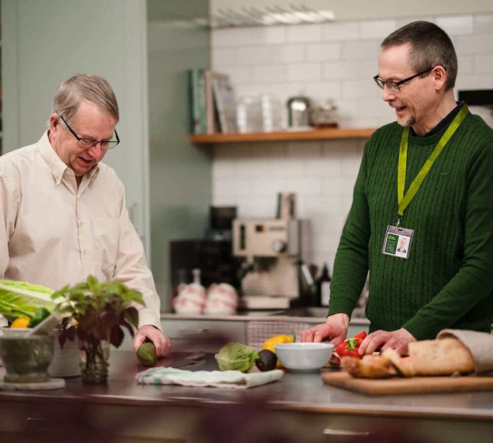 A male carer wearing green with eyeglasses happy and smiling while cuttign vegetables with an older male adult with grey hair and eyeglasses inside the kitchen both happy and smiling