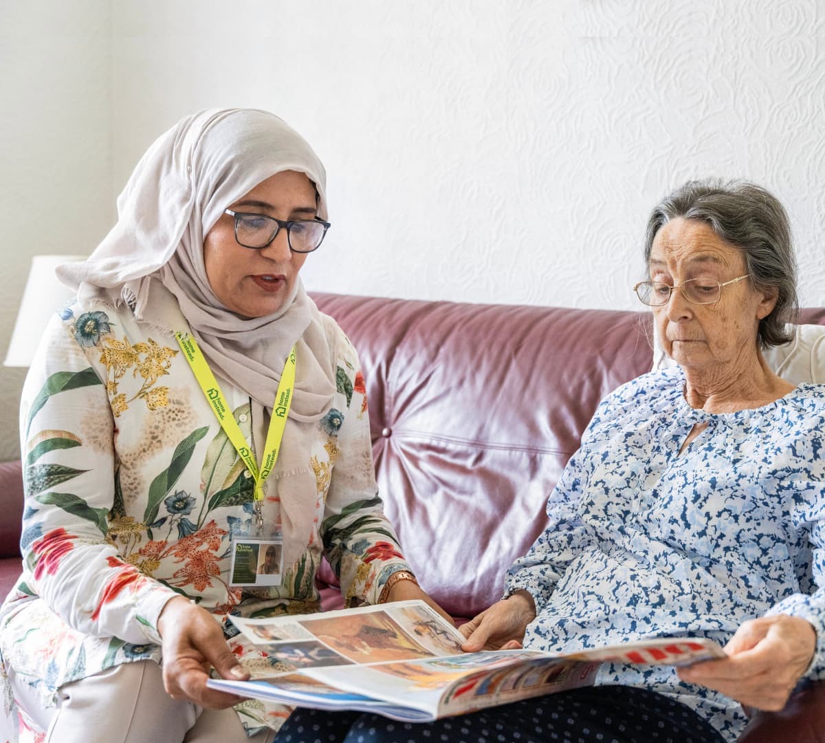 An older female adult reading the newspaper while sitting on a couch with her younger female carer wearing eyeglasses inside the house