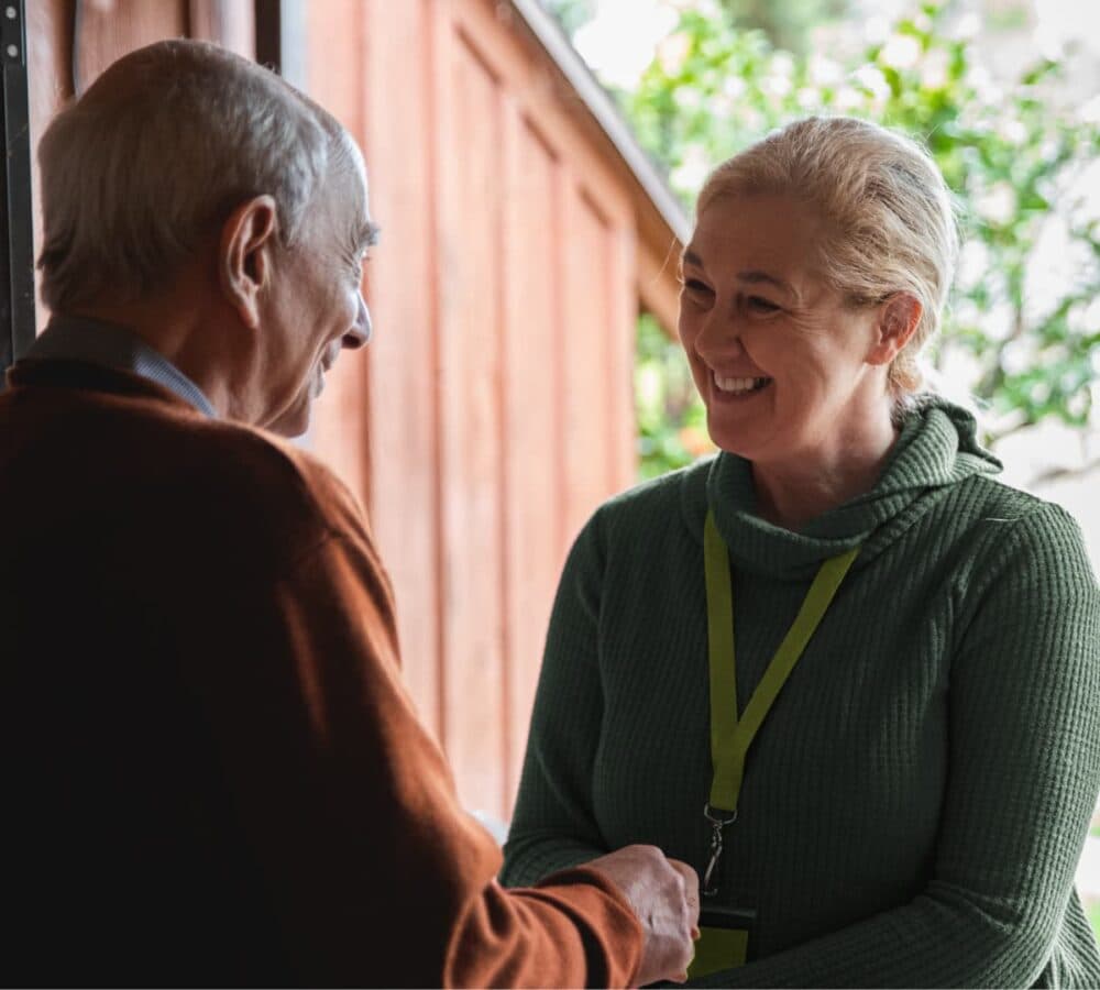 A woman at the door with blonde hair and wearing green shaking hands with an older male adult