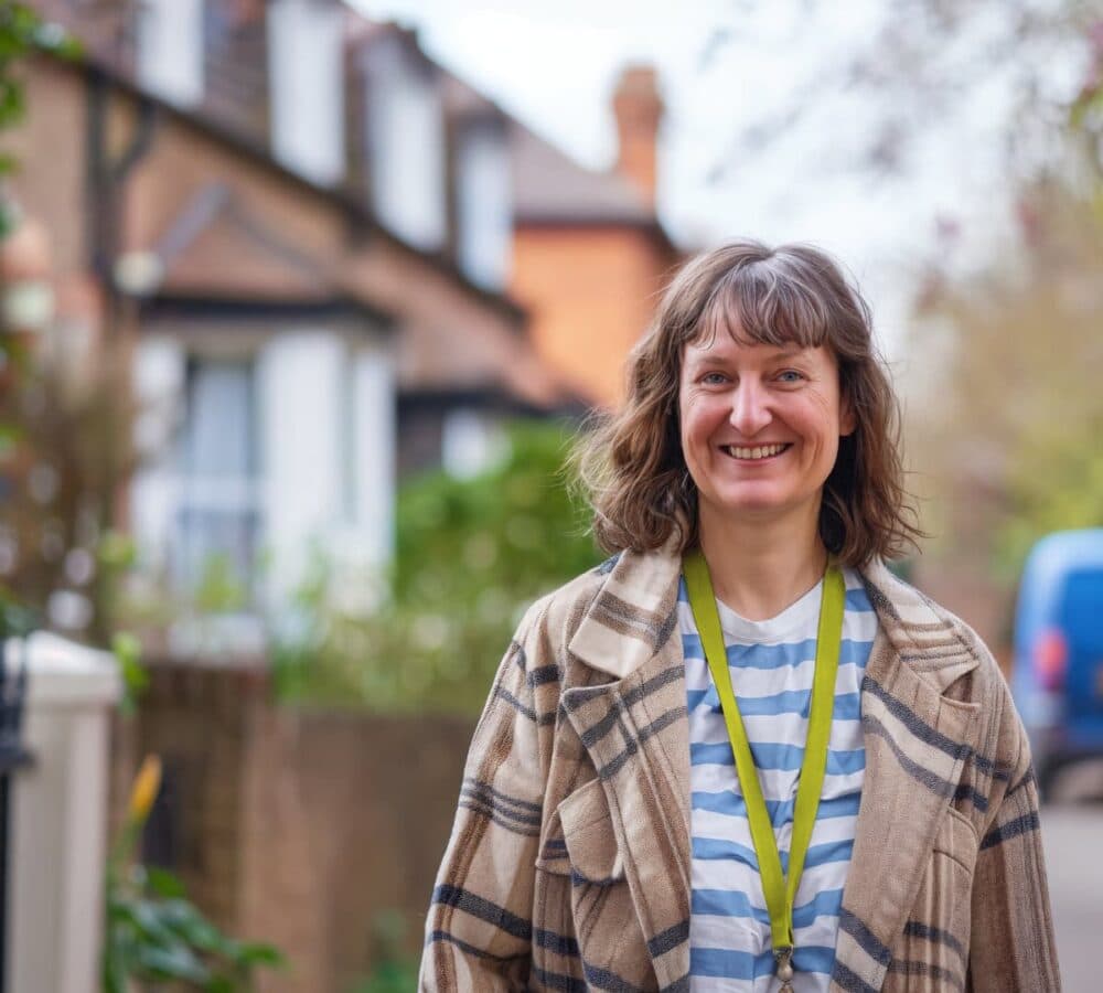 Smiling woman wearing a striped shirt and plaid coat stands outside on a residential street. - Home Instead